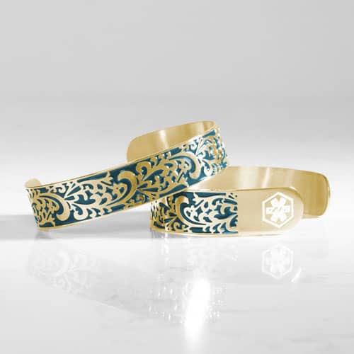 Two gold-tone medical ID cuffs featuring intricate blue filigree detailing, with one showing a white medical symbol.