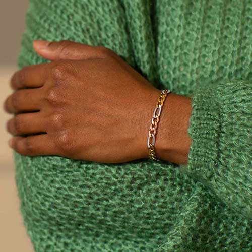Woman in green sweater wearing mixed metal stainless steel figaro chain bracelet with silver, rose gold, and gold tones.