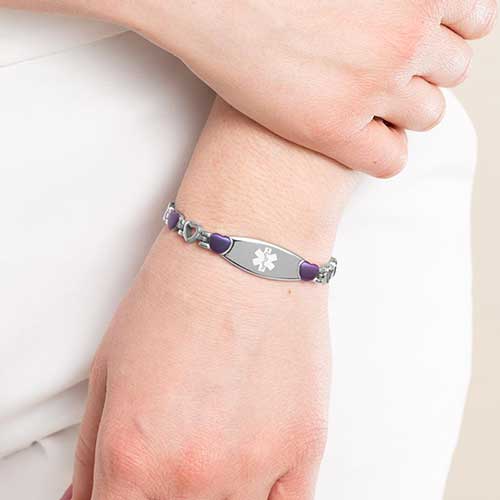 Woman wearing purple tone and and stainless steel heart link bracelet with attached medical ID tag with white medical symbol.