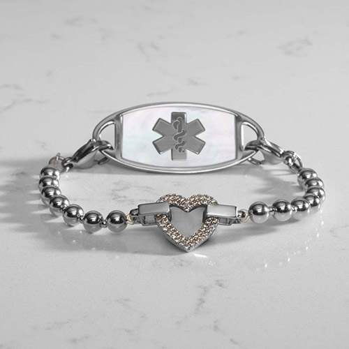 Silver medical alert bracelet with a beaded strand and a heart-shaped centerpiece encrusted with clear crystals, paired with a white mother-of-pearl medical ID tag engraved with a silver medical symbol, displayed on a white marble surface.