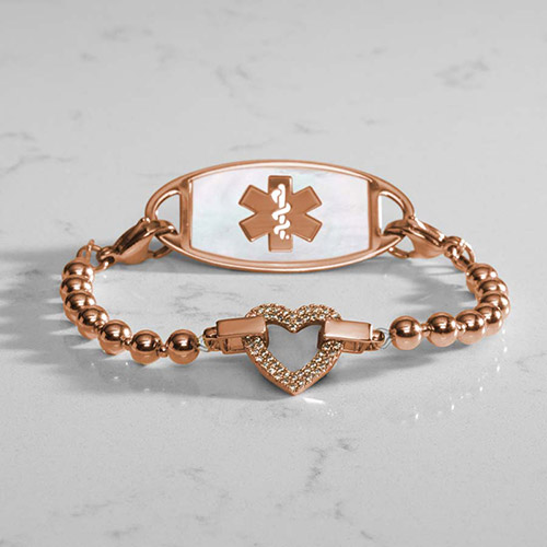 Rose gold medical alert bracelet with a beaded strand and a heart-shaped centerpiece encrusted with clear crystals, paired with a white mother-of-pearl medical ID tag engraved with a silver medical symbol, displayed on a white marble surface.