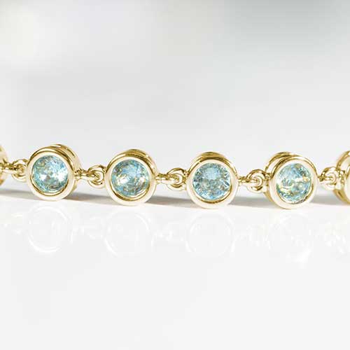 Close up image of gold tennis bracelet with individually linked, round, aquamarine cubic zirconias.