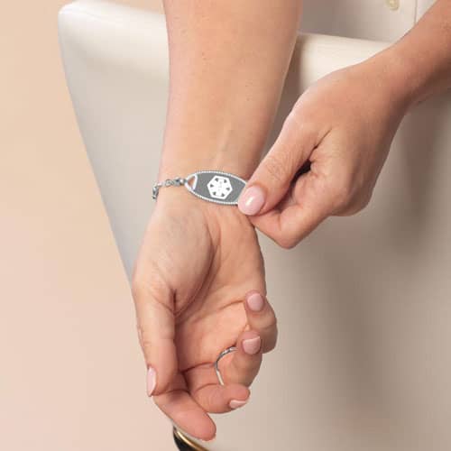 Close-up of silver medical ID bracelet with white enamel star of life symbol and delicate beaded chain on woman’s wrist