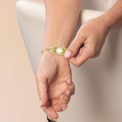 Close-up of gold medical ID bracelet with white enamel star of life symbol and delicate beaded chain on woman’s wrist