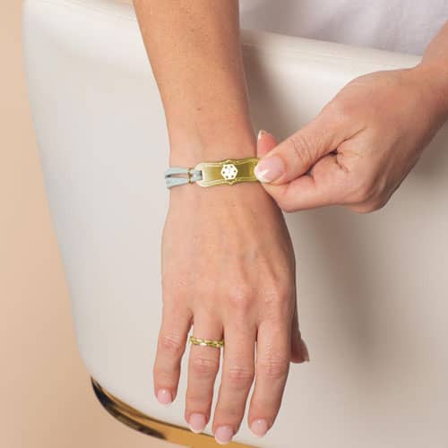 Woman wearing Avery SmartSize Medical ID Bracelet with suede light blue tone leather band and adjustable chain with a Gold La Petite tag with a white Cadeusus 