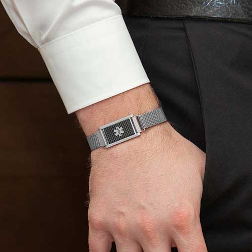 Man in white shirt wearing stainless steel mesh bracelet with magnetic closure and carbon fiber medical alert tag with hand in pocket.