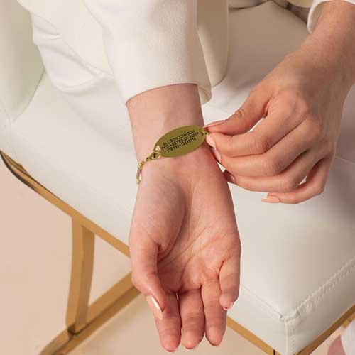 Engraved gold medical alert tag on wrist, paired with pastel beaded bracelet for style and safety.