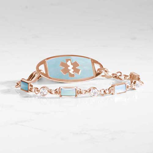 Rose medical ID bracelet with blue mother-of-pearl inlay tag and clear and iridescent rectangular gemstone accents, displayed on a marble background.