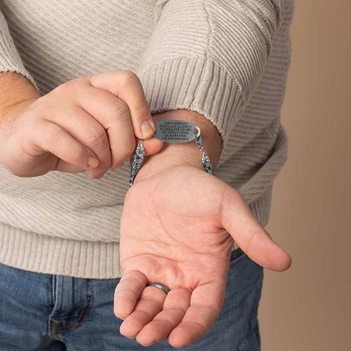 Close-up of a man’s hand adjusting a men’s medical ID bracelet with custom engraving for pacemaker, allergies, and emergency contact information, shown on a stainless steel chain.