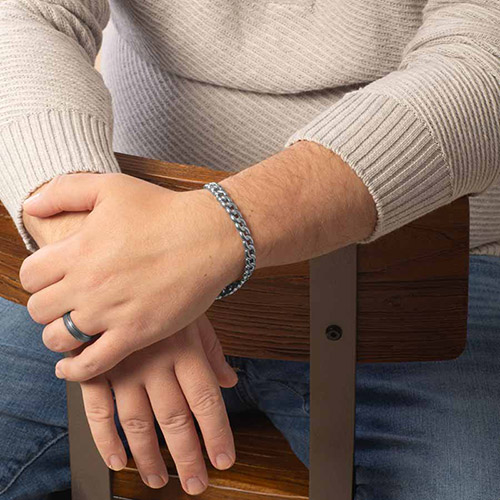 Man sitting on a wooden chair wearing a men’s medical ID bracelet with a classic silver curb chain design, styled for everyday wear.
