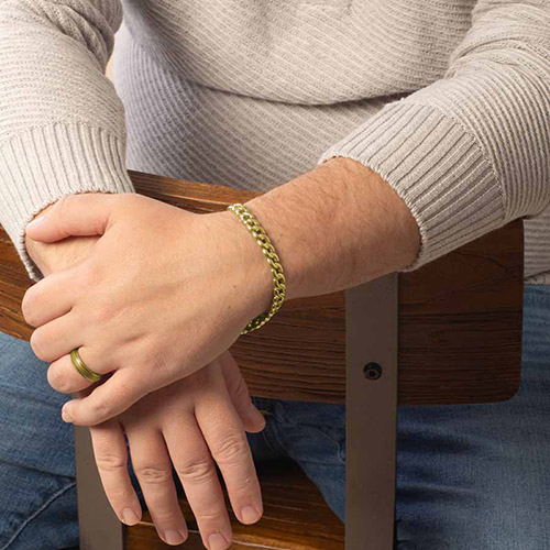Close-up of a man resting his arms on a chair while wearing a stylish gold medical alert bracelet with a flat curb chain design.