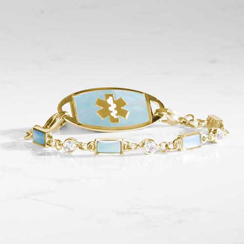 Gold medical ID bracelet with blue mother-of-pearl inlay tag and clear and iridescent rectangular gemstone accents, displayed on a marble background.