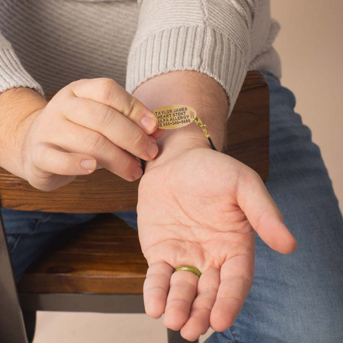 A man’s hand holding up a gold medical ID tag on the inside of his wrist, showing custom black engraving details on a black and gold bracelet.