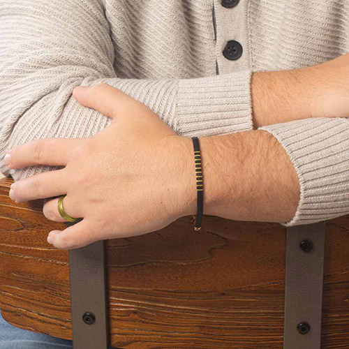 A close-up side view of the same black bracelet with gold rectangular accents, worn on the wrist with arms crossed over the back of a wooden chair.