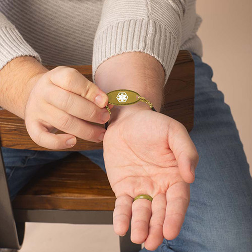 A front-facing view of the gold medical ID tag on the man’s wrist, displaying the white medical caduceus symbol on the brushed gold faceplate.