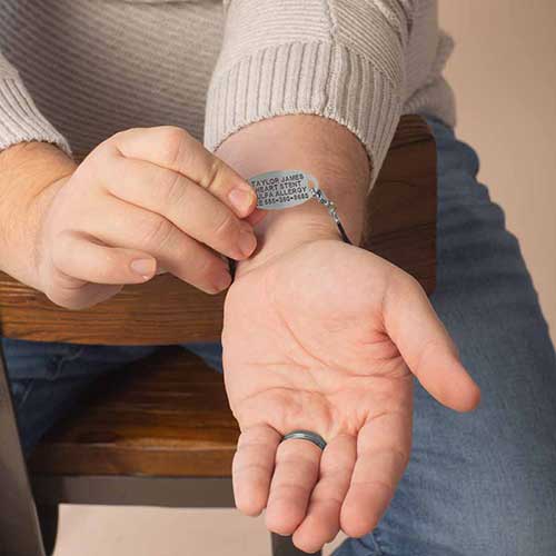 A man’s hand holding up a silver medical ID tag on the inside of his wrist, showing custom black engraving details on a black and silver bracelet.