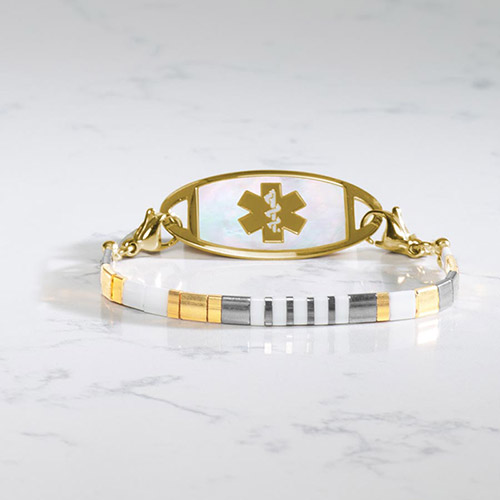 Close-up of the Nova Gold medical ID bracelet featuring a white, gold, and silver striped beaded strand paired with a mother-of-pearl ID tag and gold medical alert symbol.