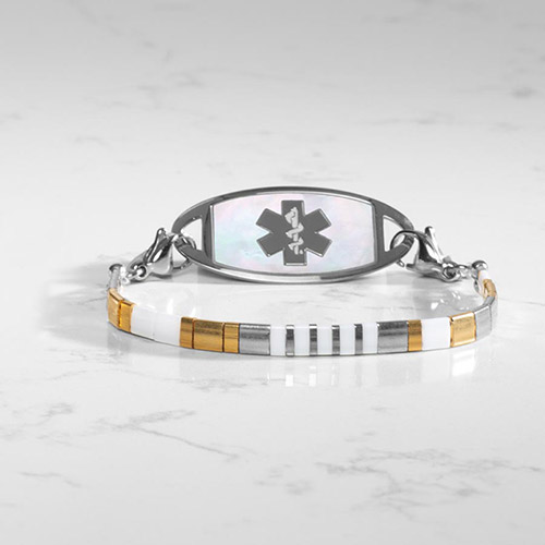 Silver medical ID bracelet with a mother-of-pearl tag, paired with a stretch strand of white, silver, gold, and hematite cube beads, shown on marble surface.