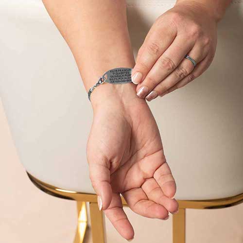 Woman’s wrist wearing a medical ID bracelet with customized engraving, featuring a sleek silver tag and a stylish link chain, shown in a close-up lifestyle view to highlight personalization and fit.