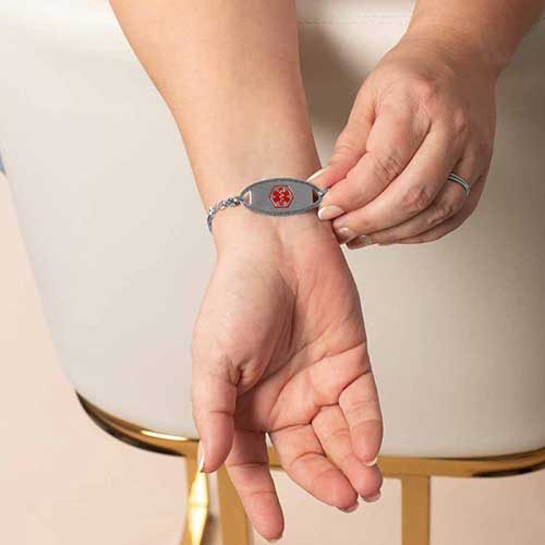 Close-up of a woman adjusting a silver medical alert bracelet on her wrist, showcasing the red medical symbol on the tag and the elegant, feminine chain design in a lifestyle setting.