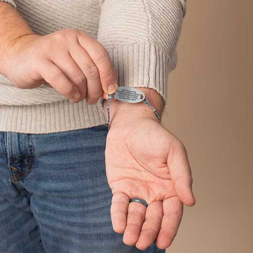Close-up of a man's wrist showing the engraved ID tag of the Mercury men's medical alert bracelet, styled with a gray shirt and wedding band.
