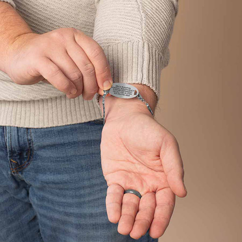 Close-up of a man's wrist showing the engraved ID tag of the Mercury men's medical alert bracelet, styled with a gray shirt and wedding band.
