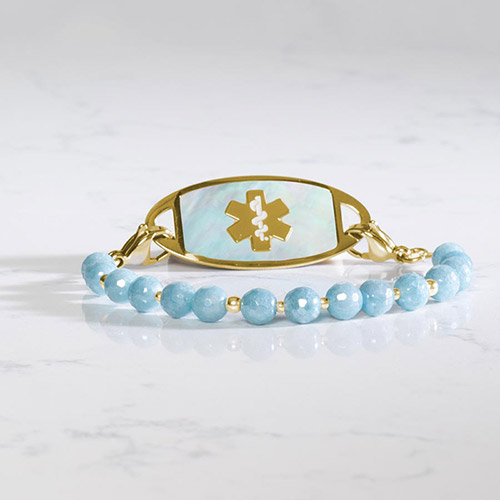 Gold medical alert bracelet with soft blue gemstone beads and a matching gold and blue ID tag, displayed on a white marble surface.