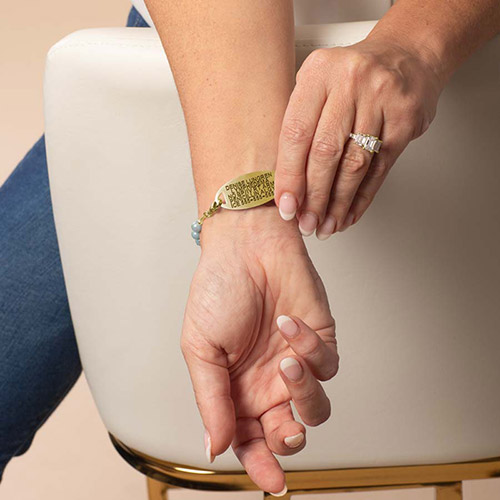 Close-up of a woman wearing a Lauren’s Hope medical ID bracelet with a gold tag and cloud blue beaded strand, engraved with emergency medical information.