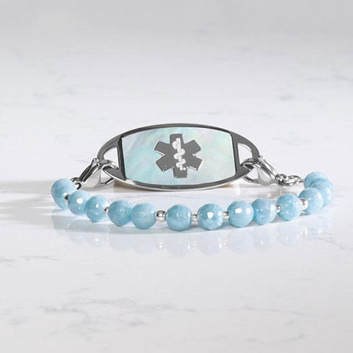 Silver medical alert bracelet resting on a marble surface, with soft lighting accentuating the glossy aquamarine beads and mother-of-pearl tag inlay.