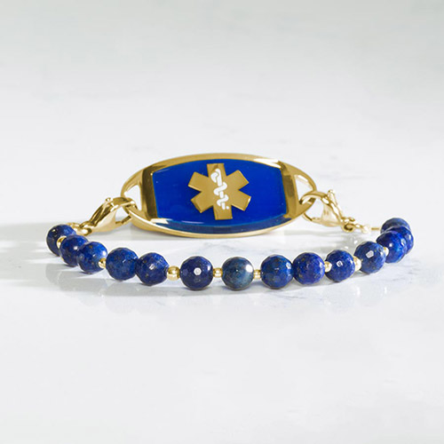 Leah bracelet in Denim shown with a gold-tone medical ID tag featuring a vibrant cobalt blue inlay and gold medical symbol, laid flat on a marble surface.