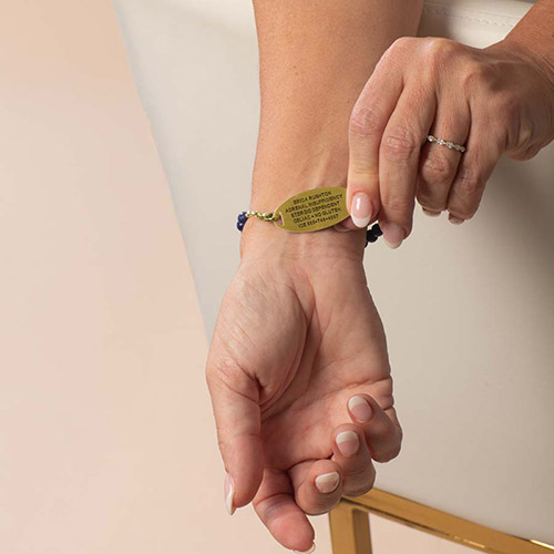 Woman’s hand turning over a gold medical ID tag to reveal custom-engraved emergency information on the back, attached to a blue beaded bracelet.