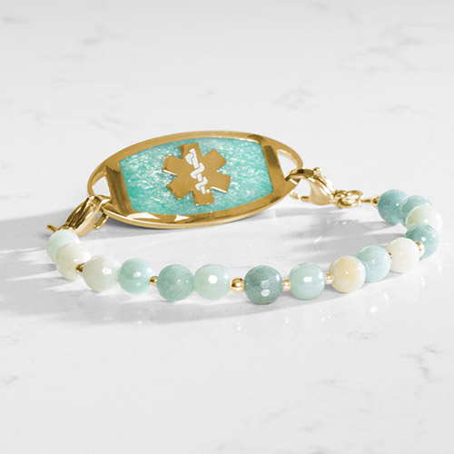 Close-up of Leah Mist medical alert bracelet in gold with soft green and ivory beads and a textured amazonite inlay tag, styled on a marble surface.