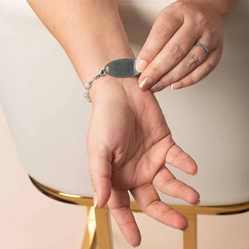 Close-up of a woman's wrist showing the underside of a medical ID bracelet with a silver engraved tag, held gently in place by her other hand. The bracelet features soft mist-colored beads and a silver chain attachment.