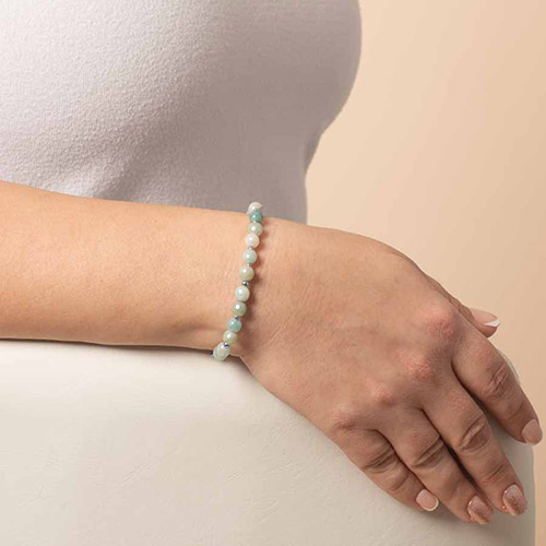 A woman’s arm resting on a white surface, wearing a delicate beaded medical ID bracelet in pale green and misty blue tones, with gold accent beads.