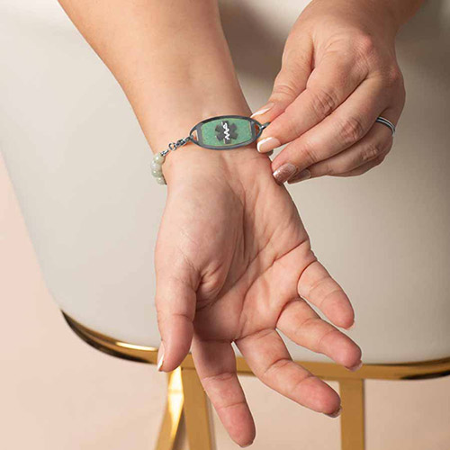 Close-up of a woman's wrist showing the front of a medical ID tag with a white medical symbol on a green background, framed in silver, attached to a beaded bracelet.