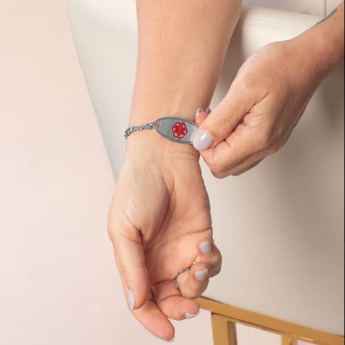 Hand lifting a silver tone medical ID tag to show the front with white inlay and medical symbol.
