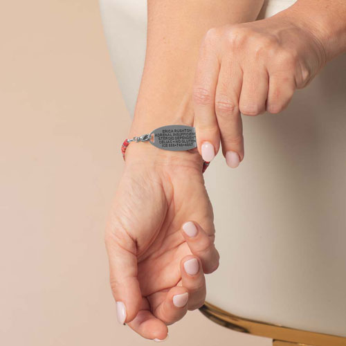 Person displaying the engraved back of a silver-tone medical ID tag attached to a pink heishi bead bracelet.