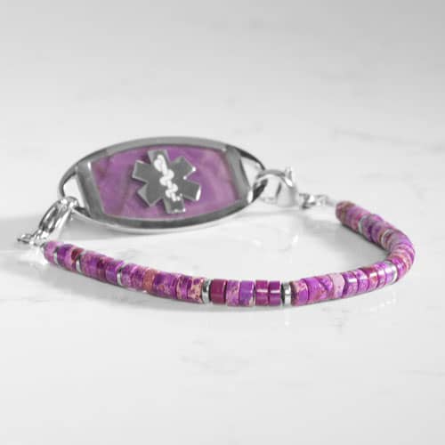 Silver-tone medical ID bracelet with purple stone inlay ID tag and purple heishi beads displayed on a white pedestal.