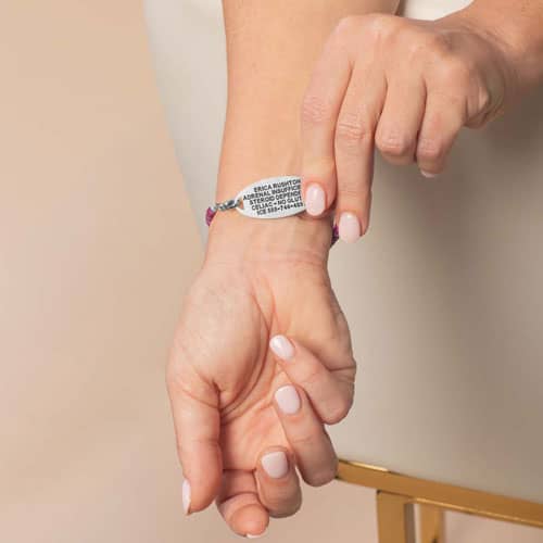 Person displaying the engraved back of a silver-tone medical ID tag attached to a purple heishi bead bracelet.