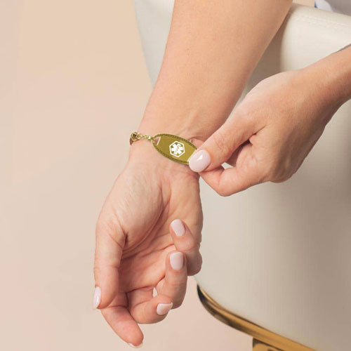Model displaying a gold-tone oval medical ID tag with a white Star of Life symbol and decorative border on the wrist.