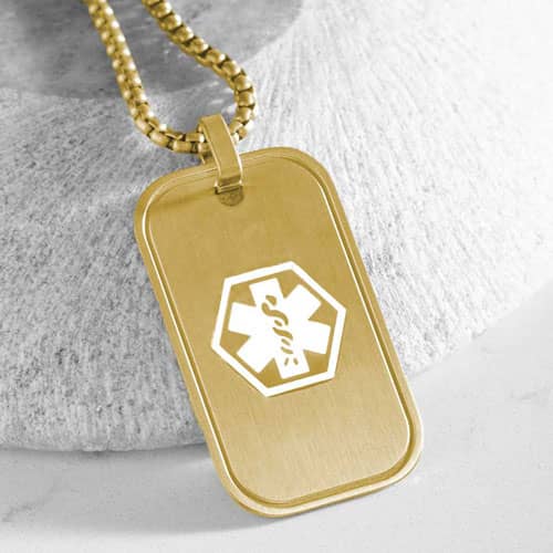 Close-up of a gold-tone medical ID dog tag necklace featuring a white medical symbol, displayed against a textured neutral background.