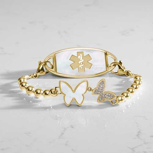 Serenity Stretch medical ID bracelet featuring gold filled beads and adouble butterfly centerpiece adorned with crystal and mother of pearl inlay, attached to a gold medical ID tag featuring mother of pearl inlay and a gold caduceus.