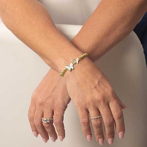 Woman displaying gold tone, beaded stretch bracelet with double butterfly centerpiece featuring crystals and mother of pearl inlay.