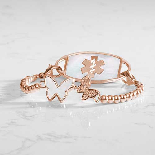 Serenity Stretch medical ID bracelet featuring rose gold filled beads and a double butterfly centerpiece adorned with crystal and mother of pearl inlay, attached to a rose gold medical ID tag featuring mother of pearl inlay and a rose gold caduceus.