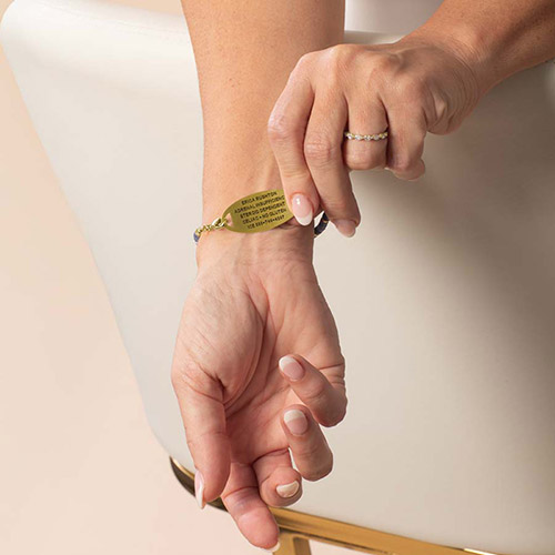 A woman lifting her wrist to show the back of the gold-tone ID tag on the Azores Gold bracelet, featuring sample medical engraving.