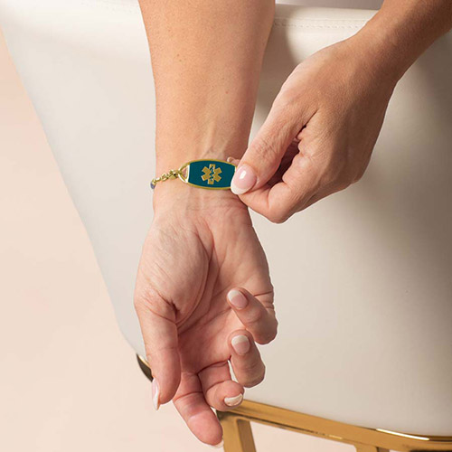 A gold-tone medical ID tag with a teal inset and engraved medical symbol, shown on the wrist as part of the Azores Gold bracelet.