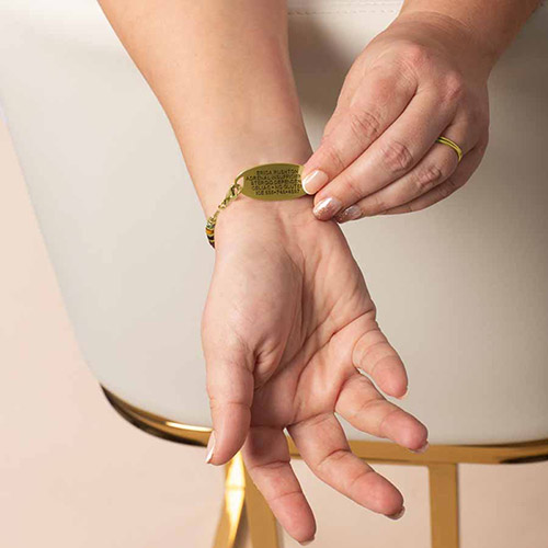 Close-up of a person’s wrist showing the underside of a gold medical ID tag engraved with emergency information, attached to a beaded bracelet in earthy green and gold tones.