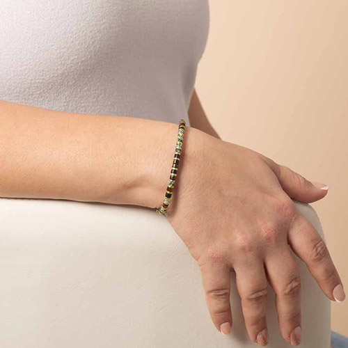 A beaded stretch bracelet in a mix of green, black, and gold colors worn on the wrist, styled with a simple white top.