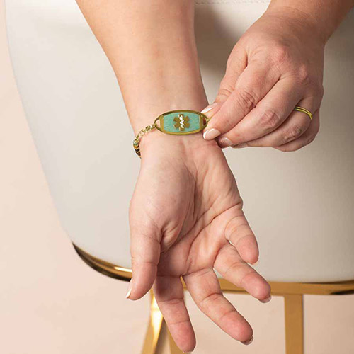 Close-up of a gold medical ID tag with a turquoise inlay and white medical symbol, shown on the wrist and paired with the green and gold beaded Ember bracelet.