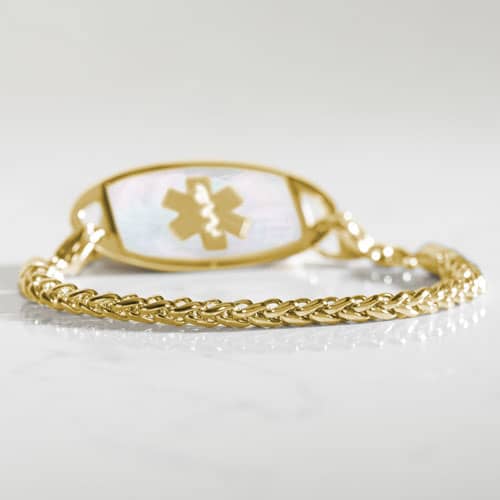 Gold-tone wheat chain medical ID bracelet with mother-of-pearl tag photographed on a reflective surface.